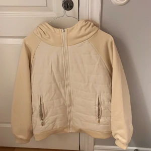 Beige quiltad jacka med huva - Säljer en snygg beige jacka med quiltad design och dragkedja framtill. Jackan har en huva och två praktiska fickor med dragkedjor. Perfekt för kyligare dagar!