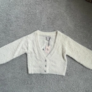 Vit fluffig kofta från Sincerely Jules - Säljer en supermjuk och fluffig vit kofta från Sincerely Jules. Den har en v-ringad design med knappar framtill och är croppad i modellen. Helt ny och oanvänd med prislapp på!