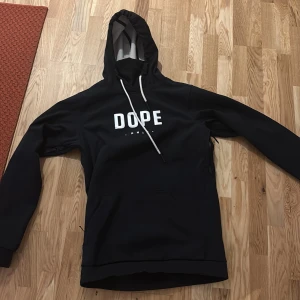 Svart skidjacka från Dope - Säljer en svart hoodie från Dope med vit text på bröstet. Den har en justerbar huva med vita snören och långa ärmar. Perfekt för en avslappnad stil