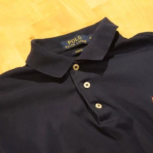 Mörkblå pikétröja från Ralph Lauren - Säljer en klassisk mörkblå pikétröja från Ralph Lauren. Tröjan har en stilren design med krage och tre knappar framtill. Perfekt för en avslappnad men ändå elegant look.