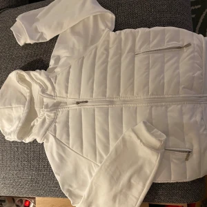 Vit cardigan jacka med huva - Säljer en stilren vit cardigan jacka med huva. Jackan har dragkedja framtill och två praktiska fickor med dragkedjor. Perfekt för mildare dagar med sina långa ärmar. Storleken är small