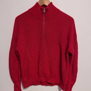 Röd stickad tröja från JJXX - Snygg röd stickad tröja från JJXX med dragkedja i halsen. Perfekt för kyligare dagar med sin varma och bekväma design. Tröjan har långa ärmar och en ribbad struktur som ger en stilren look.