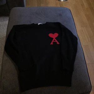 Svart tröja från Ami Alexandre Mattiussi - Snygg svart tröja från Ami Alexandre Mattiussi med ett rött hjärta och bokstaven 'A' på bröstet. Tröjan har långa ärmar och en klassisk rund halsringning. Perfekt för en stilren look.
