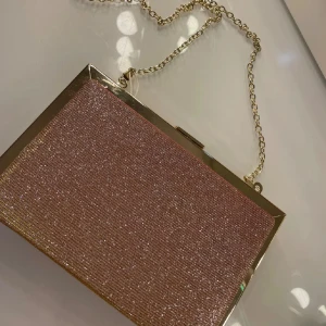 Glittrig rosa kuvertväska med kedja - Snygg glittrig rosa kuvertväska med en elegant guldfärgad kedja. Perfekt för att ge en touch av glamour till din outfit. Väskan har en stilren design med en hård ram och en praktisk stängning.