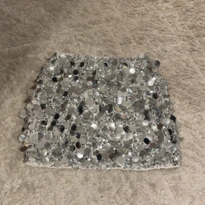 Glittrig silverkjol med paljetter - Snygg och iögonfallande kortkjol i silver täckt med stora paljetter. Perfekt för att sticka ut på festen. Kjolen har en tight passform och är gjord i ett syntetmaterial som ger en glittrig effekt.