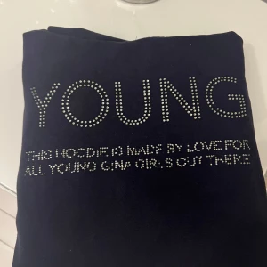 Marin blå  hoodie med textdetalj - Säljer en marin blå hoodie med texten 'YOUNG' i glittriga stenar på framsidan. Hoodien har långa ärmar och en klassisk huva. Perfekt för en avslappnad stil.