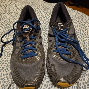 Svarta och blåa löparskor från Asics - Säljer ett par Asics löparskor i svart och blått med FlyteFoam-sula för extra komfort. Skorna har snörning och är tillverkade i syntetmaterial med gummisula. Perfekta för löpning och träning.