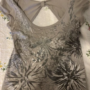 Grå/Beige blommig topp från New Look - Säljer en beige/grå topp från New Look med blommigt mönster och glittriga detaljer. Toppen har korta ärmar och en öppen rygg med knytning. Perfekt för en stilfull look. Står inte storlek men skulle gissa på att den är S.💗