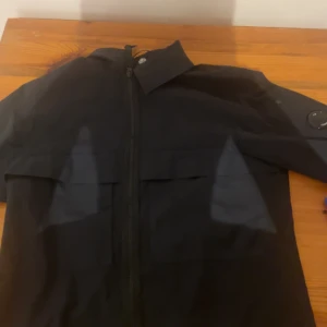 Svart jacka från C.P. Company - Snygg svart jacka från C.P. Company med dragkedja och coola detaljer. Perfekt för en stilren look. Märkeslogga på ärmen ger en extra touch av stil.