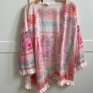 Tunika  med fransar s 152 - Säljer en färgglad tunika med blommigt mönster i rosa, aprikos och grönt. Den har fransdetaljer längs nederkanten.