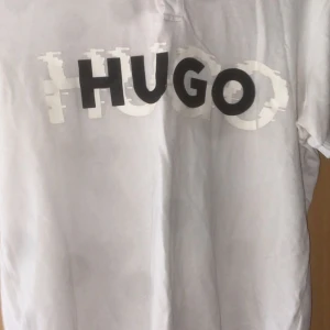 Vit t-shirt från Hugo - Säljer en stilren vit t-shirt från Hugo med stort tryck av märkesnamnet i svart och vitt på framsidan. T-shirten har en klassisk passform och korta ärmar, perfekt för en avslappnad look.