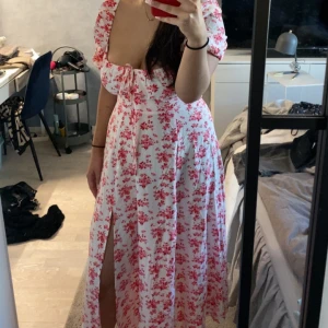 Blommig långklänning med slits - Charmig vit långklänning med rosa blommönster och en djup v-ringning. Klänningen har korta ärmar och en elegant slits framtill. Perfekt för somriga dagar och speciella tillfällen.