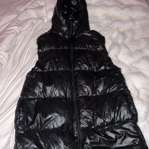 Svart dunväst från H&M - Säljer en svart dunväst från H&M med dragkedja och huva. Perfekt för kyliga dagar när du vill hålla dig varm och bekväm. Västen har en glansig yta som ger en modern look.