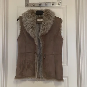 Beige pälsväst från Intown - Säljer en snygg beige pälsväst från Intown med mjuk fuskpäls längs kragen och kanterna. Västen har en stilren design och är perfekt för att hålla sig varm och trendig. I storlek 36 o kan prutas 