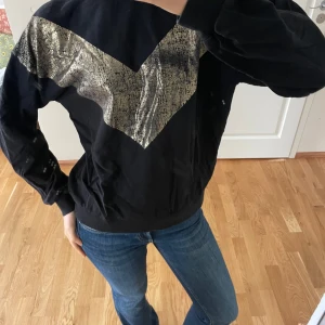 Svart tröja med glittrig detalj - Snygg svart tröja med en glittrig silverdetalj i ett V-format mönster över bröstet. Tröjan har långa ärmar och en avslappnad passform, perfekt för en trendig look.