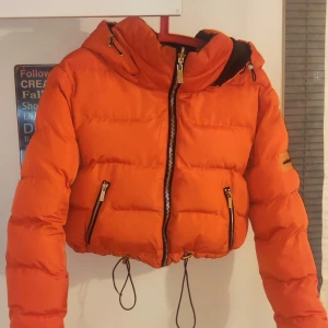 Orange och svart pufferjacka - Säljer en cool och vändbar pufferjacka i orange och svart. Jackan har en dragkedja framtill och justerbara snören i nederkant. Perfekt för att hålla värmen med stil!