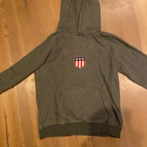 Grå hoodie från GANT - Säljer en grå hoodie från GANT med broderad logga och ett emblem med stjärnor och ränder på framsidan. Tröjan har en klassisk känguruficka och ribbade muddar. Perfekt för en avslappnad stil. Ord pris 699kr