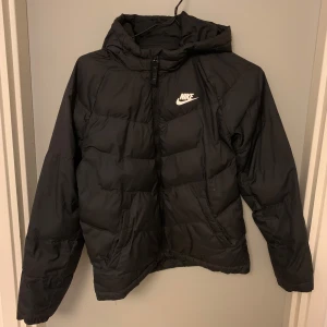 Svart dunjacka från Nike - Säljer en svart dunjacka från Nike med huva och dragkedja. Jackan har en klassisk pufferdesign och är perfekt för kyligare väder. Nike-loggan är tryckt på bröstet för en sportig look.