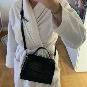 Svart quiltad handväska - Snygg svart handväska med quiltat mönster och avtagbar axelrem. Väskan har ett stilrent lock med magnetknäppning och ett rymligt inre med dragkedjefack. Perfekt för att bära dina nödvändigheter med stil.