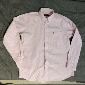 Rosa och vit randig skjorta från Polo Ralph Lauren - Snygg rosa och vit randig skjorta från Polo Ralph Lauren med broderad logga på bröstet. Skjortan har långa ärmar och knappar framtill. Perfekt för en stilren look!