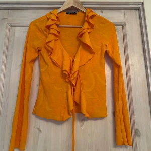 Orange volangtröja från Gina Tricot - Säljer en snygg orange tröja från Gina Tricot med volangdetaljer och knyt framtill. Tröjan har långa ärmar och en djup v-ringning, perfekt för att ge en färgklick till din outfit. Passar bra till både jeans och kjol. 