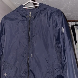 Moncler vindjacka - Snygg mörkblå vindjacka från Moncler med dragkedja och huva. Jackan har Moncler-logga på ärmen och röd-vit-blå randdetalj på huvan. 