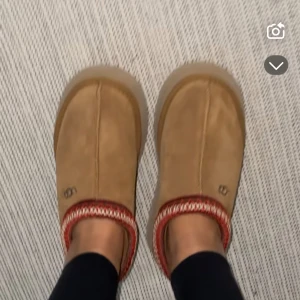UGG TAZZ CHESTNUT - Snygga bruna slip-ons i mocka med en dekorativ röd och vit mönstrad kant. Perfekta för en avslappnad stil. De har en bekväm sula och är lätta att ta på och av.