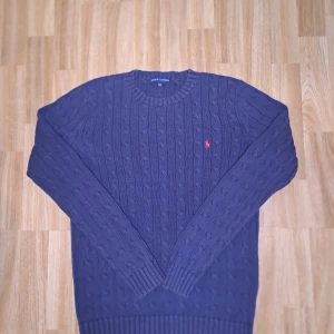 Mörkblå stickad tröja från Ralph Lauren - Säljer en snygg mörkblå stickad tröja från Ralph Lauren med kabelstickat mönster. Tröjan har en broderad röd logotyp på bröstet och långa ärmar. Perfekt för kyliga dagar!