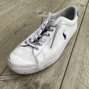 Polo Ralph Lauren Skor - Snygga vita sneakers från Polo Ralph Lauren med klassisk design och blå logotyp på sidan. Skorna har snörning och är tillverkade i skinn. De är storlek 42 men funkar för 43 tycker jag. Köpte de för 1600 men släpper för 800. Skriv vid frågor