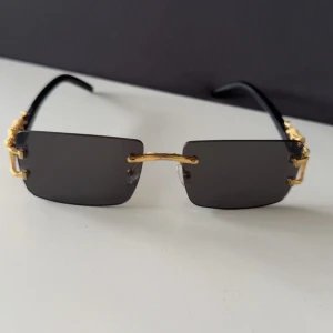 Svarta rektangulära solglasögon med gulddetaljer - Snygga solglasögon med rektangulära svarta linser och guldfärgade detaljer. Bågarna är dekorerade med unika gulddetaljer som ger en lyxig känsla. Perfekta inför sommaren. Använd 2 gånger
