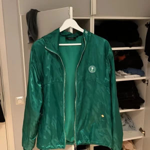 Grön vindjacka från Trapstar - Säljer en grön vindjacka från Trapstar med dragkedja och en cool logga på bröstet. Jackan har långa ärmar och en stilren design som passar perfekt för vår och höst. Den är tillverkad i ett lätt syntetmaterial.