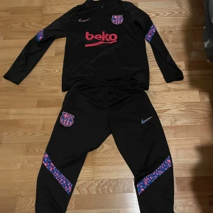 Barca  långärmad träningströja från Nike - Säljer en svart långärmad träningströja från Nike med lila och rosa detaljer. Tröjan har klubbmärket på bröstet och är perfekt för fotbollsträning. Den är tillverkad i ett syntetmaterial för optimal komfort och prestanda.