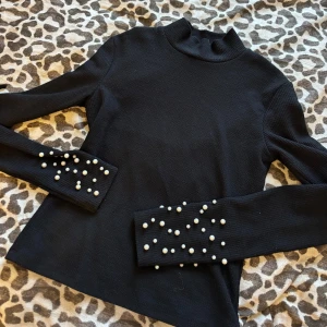 Svart ribbad polo tröja med pärlor - Snygg svart ribbad tröja med långa ärmar och dekorativa pärlor på ärmarna. Perfekt för att ge en elegant touch till din outfit. Tröjan har en hög krage och är i ett stretchigt material. (Aldrig använt)
