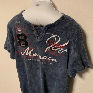 Blå t-shirt med tryck och knappar - Säljer en blå t-shirt med tryck och knappar vid halsen. T-shirten har korta ärmar och ett coolt tryck med text och siffror i rött och vitt på framsidan. Perfekt för en avslappnad stil. Storlek L.