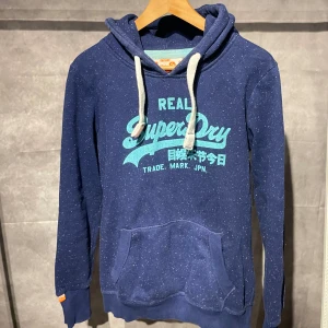 Superdry hoodie - Säljer en snygg blå hoodie från Superdry med ljusblå text och japanska tecken på framsidan. Den har en känguruficka och justerbar huva med vita snören. Perfekt för en avslappnad stil.
