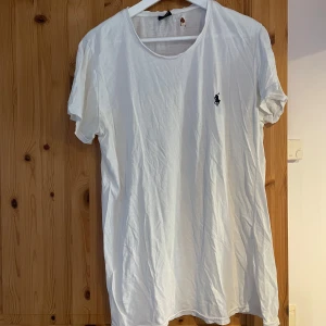 Vit t-shirt från Ralph Lauren - Säljer en klassisk vit t-shirt från Ralph Lauren med den ikoniska loggan broderad på bröstet. T-shirten har en rund halsringning och korta ärmar. Perfekt för en avslappnad stil. Sliten i nacken