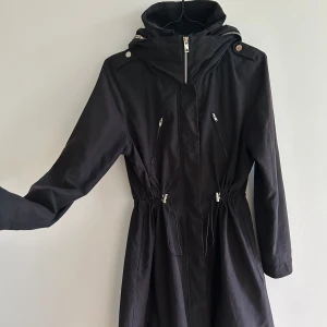 Svart kvinnlig parkajacka från Elvine - Snygg svart parkajacka i feminin modell med dragkedja och överslå med knappar. Jackan har en justerbar midja och en praktisk huva som går att rulla in i kragen när man inte vill anvånda den. Muddar i ärmarna och ett något vattensvstötande material. Perfekt för kyligare dagar med sin långa design och stilrena look. Längd till halva låret. 