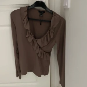 Brun omlottopp med volangdetaljer - Säljer en stilren brun omlottopp  med volangdetaljer längs halsringningen och framsidan. Toppen har långa ärmar och en elegant design som passar perfekt för höst och vår.