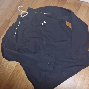 Svart vindjacka från Under Armour - Säljer en svart vindjacka från Under Armour med dragkedja och långärmade ärmar. Jackan har en stilren design med logotyp på bröstet och text på ryggen. Perfekt för träning eller en aktiv dag ute. Den är öppen vid ryggen så luft kommer in inget som är sönder. Storlek S men kan passa M beroende på bredden.