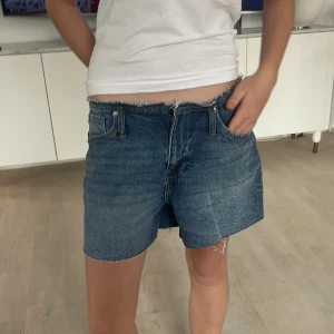 Blå jeansshorts - Säljer ett par klassiska blå jeansshorts med fransig kant. De har en normal passform och är perfekta för en avslappnad stil. Shortsen har fickor både fram och bak samt en dragkedja framtill.