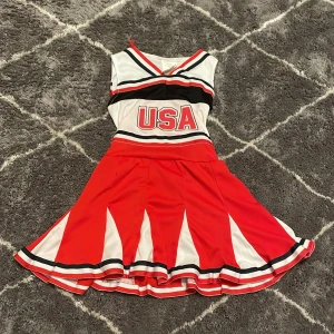 Röd och vit cheerleaderklänning med USA-tryck - Snygg cheerleaderklänning i rött och vitt med svart detaljering och USA-tryck på framsidan. Klänningen har en knytning i ryggen och är ärmlös med en v-ringad hals. Perfekt för att heja på ditt lag eller halloween disco!