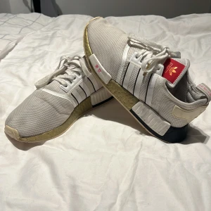Adidas NMD sneakers i vitt och beige - Snygga Adidas NMD sneakers i vitt med beige och svarta detaljer. Skorna har den klassiska tre-randiga designen och snörning framtill. Perfekta för en stilren look med en touch av streetwear.