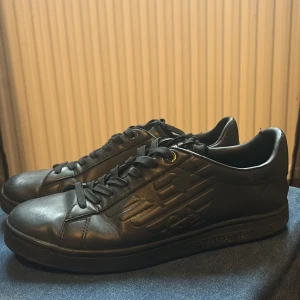 Svarta sneakers från Emporio Armani - Snygga svarta sneakers från Emporio Armani i skinn med diskret logga på sidan. Skorna har klassisk snörning och en stilren design som passar perfekt till vardags. Perfekta för den som vill ha en stilren look.Ny pris 2000kr