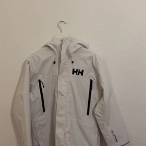 Vit vind/höst/vår jacka från Helly Hansen - Säljer en vit oanvänd jacka från Helly Hansen med dragkedja och justerbar huva. Jackan har svarta detaljer och är vattentät, perfekt för blåsiga dagar. Den har två framfickor med dragkedjor och Helly Hansen-logga på bröstet och ärmen.