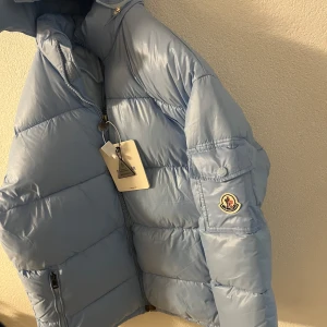 Ljusblå dunjacka från Moncler XS/S - Säljer en snygg ljusblå MAYA dunjacka från Moncler. Jackan har en dragkedja framtill och en hög krage för extra värme. Perfekt för kyliga dagar och ger en stilren look. Moncler-loggan syns på ärmen.