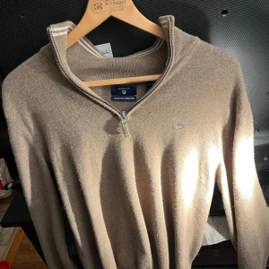 Brun ulltröja från Gant - Säljer en stilren brun tröja från Gant i superfin lammull. Tröjan har en dragkedja vid halsen och ett diskret broderat emblem på bröstet. Perfekt för kyligare dagar. Finns fler bilder vid förfrågan!