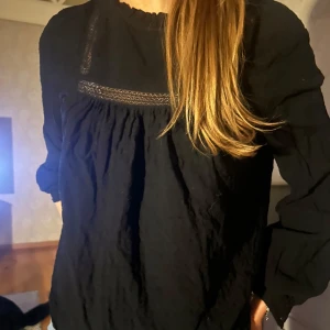 Svart blus med spetsdetaljer - Elegant svart blus med spetsdetaljer vid halsen. Blusen har långa ärmar och en lös passform, perfekt för en stilren look.