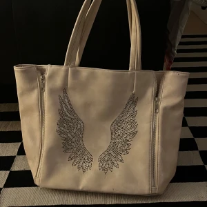 Beige axelväska med vingdetaljer - Snygg beige axelväska med glittriga vingdetaljer på framsidan. Väskan är rymlig, har bara inte använts något därav säljs👍🏼💕