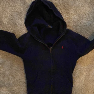 Mörkblå hoodie från Polo Ralph Lauren - Säljer en snygg mörkblå hoodie från Polo Ralph Lauren med dragkedja och broderad röd logga på bröstet. Perfekt för en casual look med sin bekväma passform och långa ärmar. Huvan ger extra stil och värme.