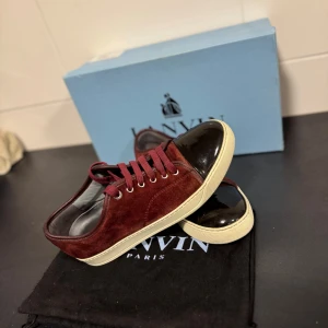 Vinröda sneakers med svart tå - Snygga vinröda sneakers från Lanvin med svart, glansig tå och vit sula. Skorna har snörning och är tillverkade i mocka vilket ger en lyxig känsla. Perfekta för att ge din outfit en stilfull touch. Box och lanvin påse följer med✅